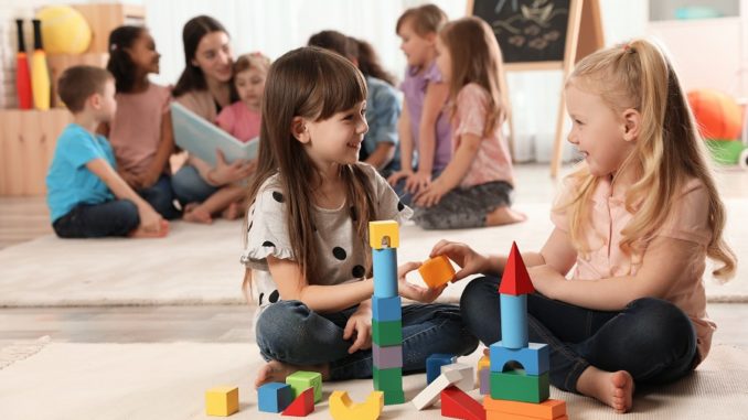 Cute girls playing with building blocks on floor while kindergarten teacher reading book to other children indoors